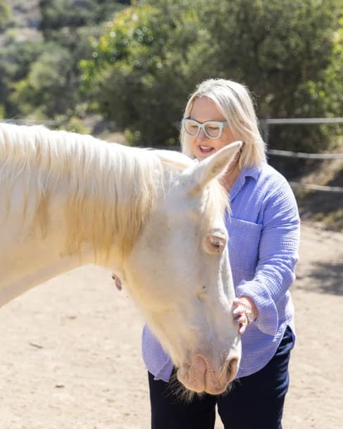 Angela Schellenberg in a lilac shirt, one hand under the jaw of a cremello horse