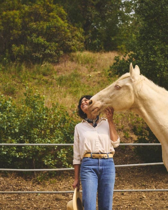 Eloisa Ribeiro laughing, hat in hand, as a cremello horse leans in to her
