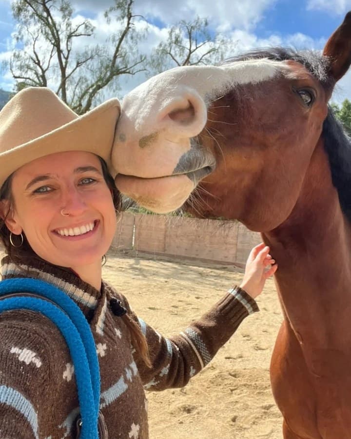 Emma Barnosky in a felt hat, laughing, as a bay horse leans into her shoulder